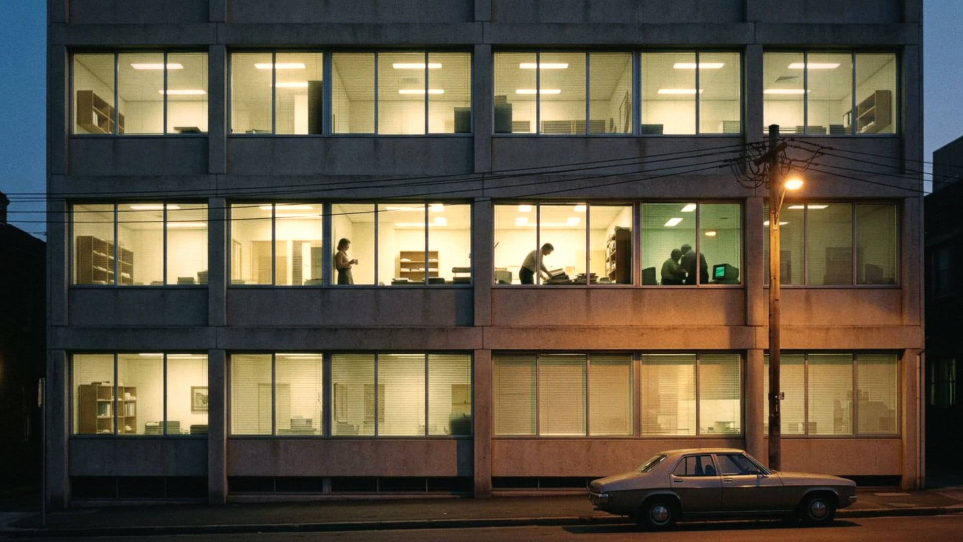 A 1970s office block on a Sydney street at dusk, people at work in the lit windows seen only in silhouette, a terminal glowing green on the late shift.